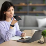 Une femme assise à un bureau, tenant un smartphone et enregistrant un message vocal, avec un ordinateur portable, un carnet, et des accessoires de bureau en arrière-plan.