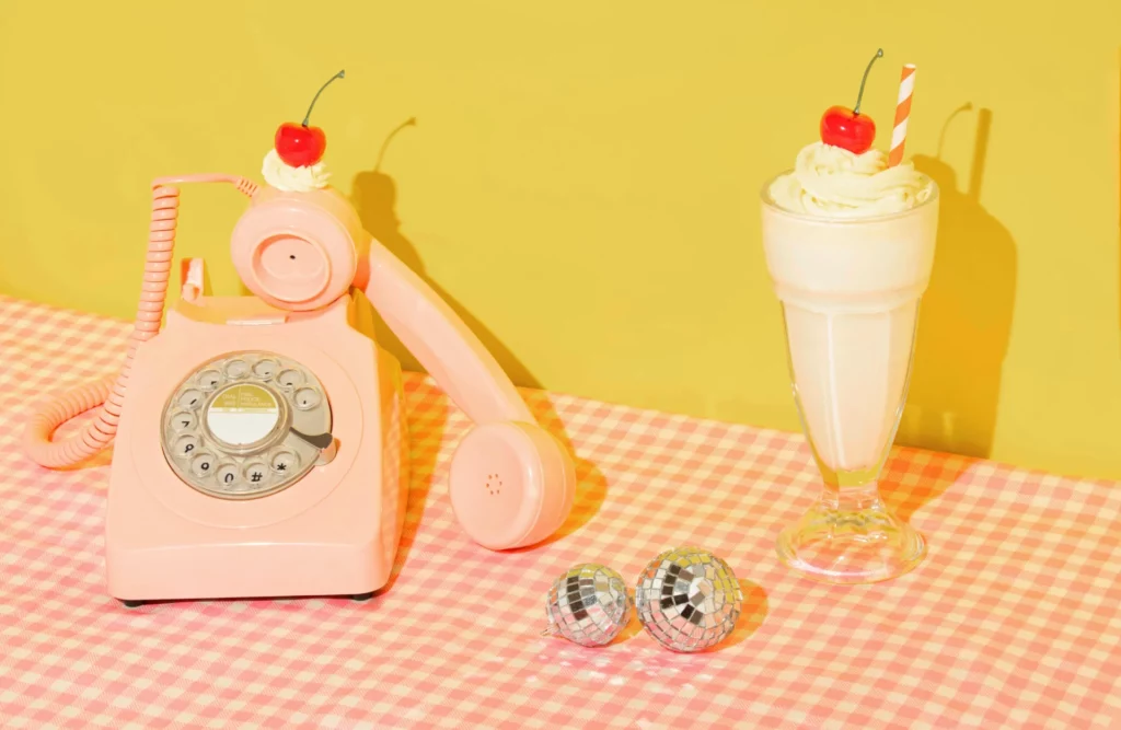 Téléphone rose vintage à côté d'un milkshake à la cerise sur une table à carreaux roses et blancs.